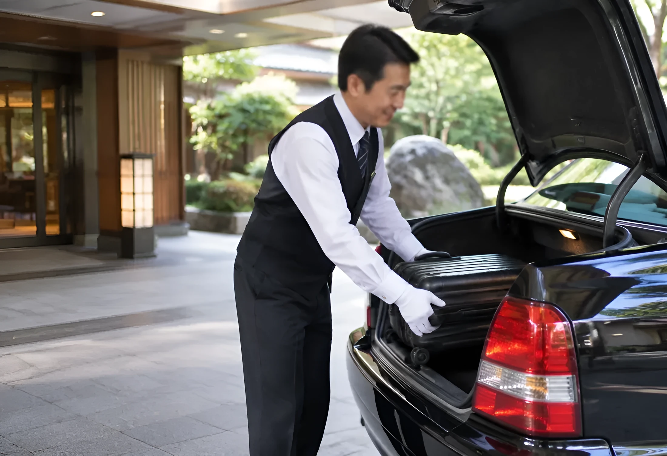 建物の車寄せで、黒い車のトランクに荷物を積む運転手