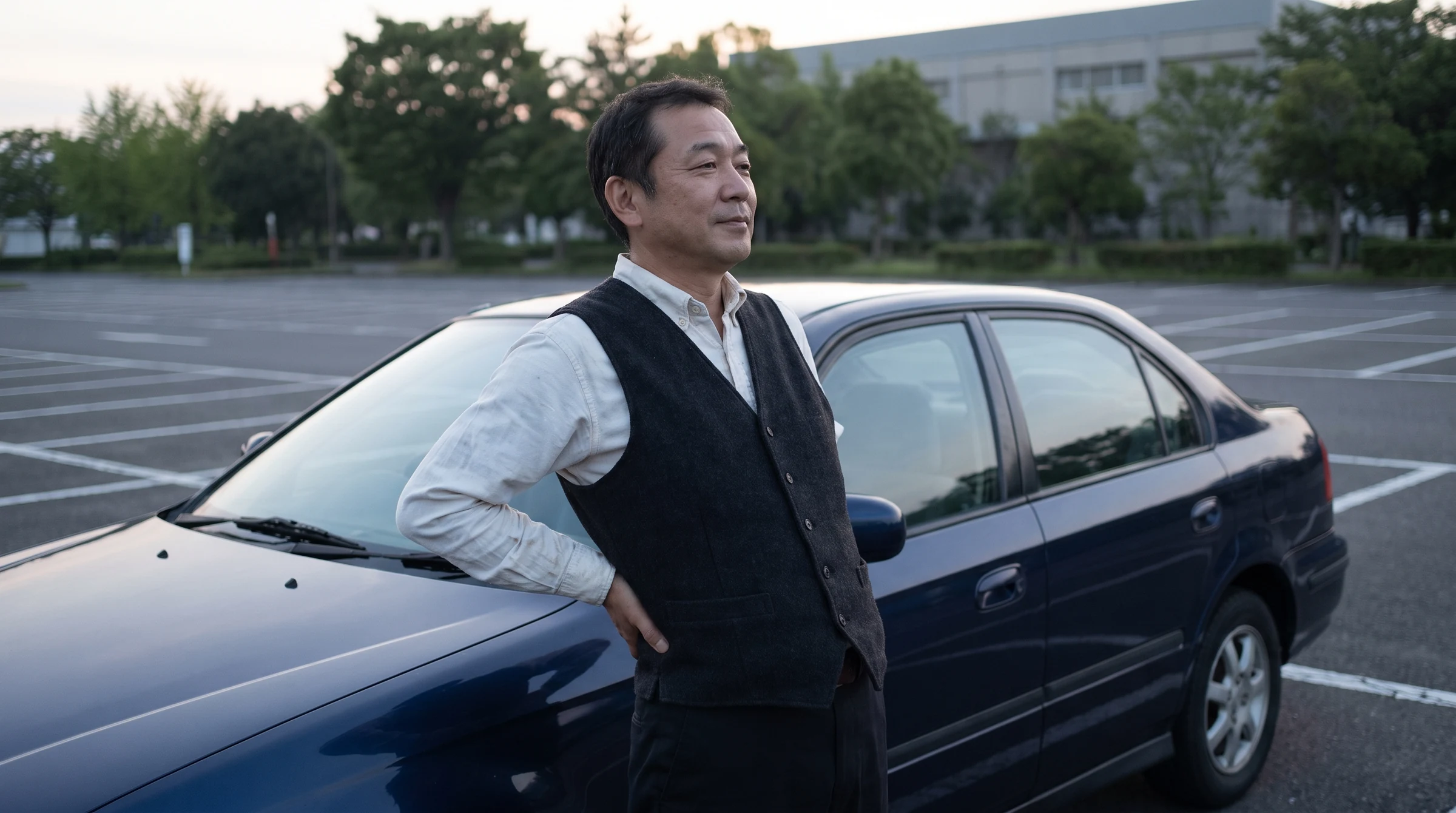 駐車場の車の横に立ち、腰に手を当てている男性
